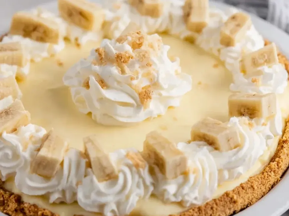 A whole pie with a golden graham crumb crust, smooth pale yellow cream filling, a ring of piped whipped cream topped with banana chunks and a centre swirl with crumbs