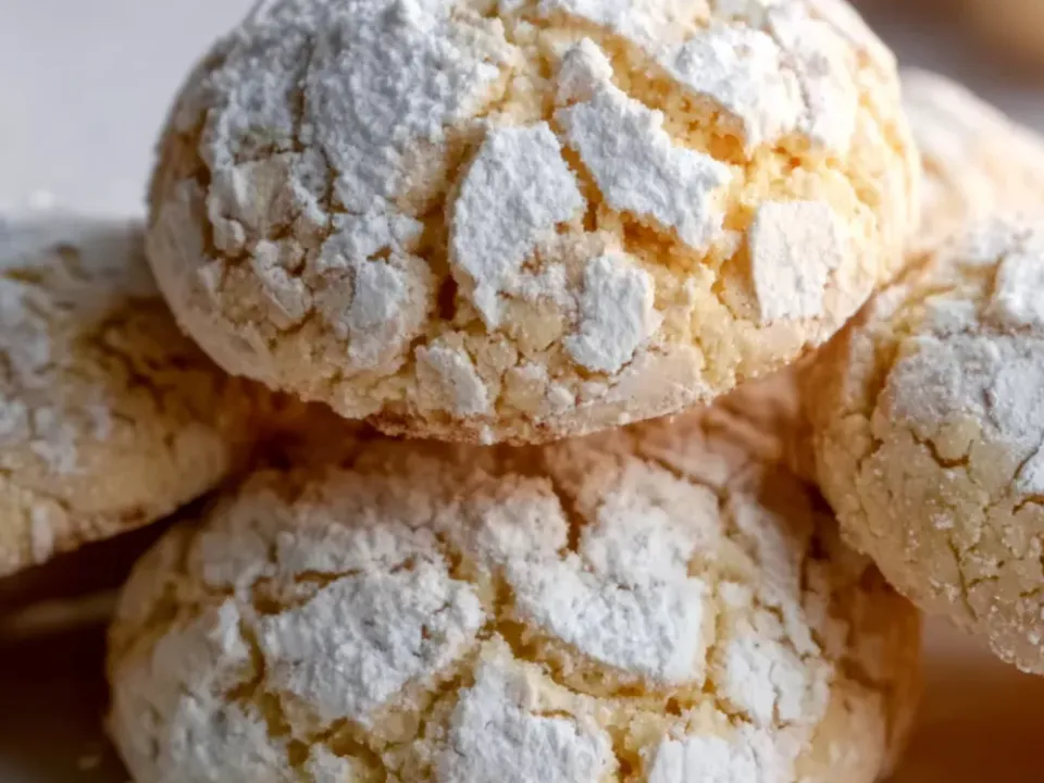 Domed cookies with deep cracks through a thick powdered-sugar coating, pale inside, piled on a plate