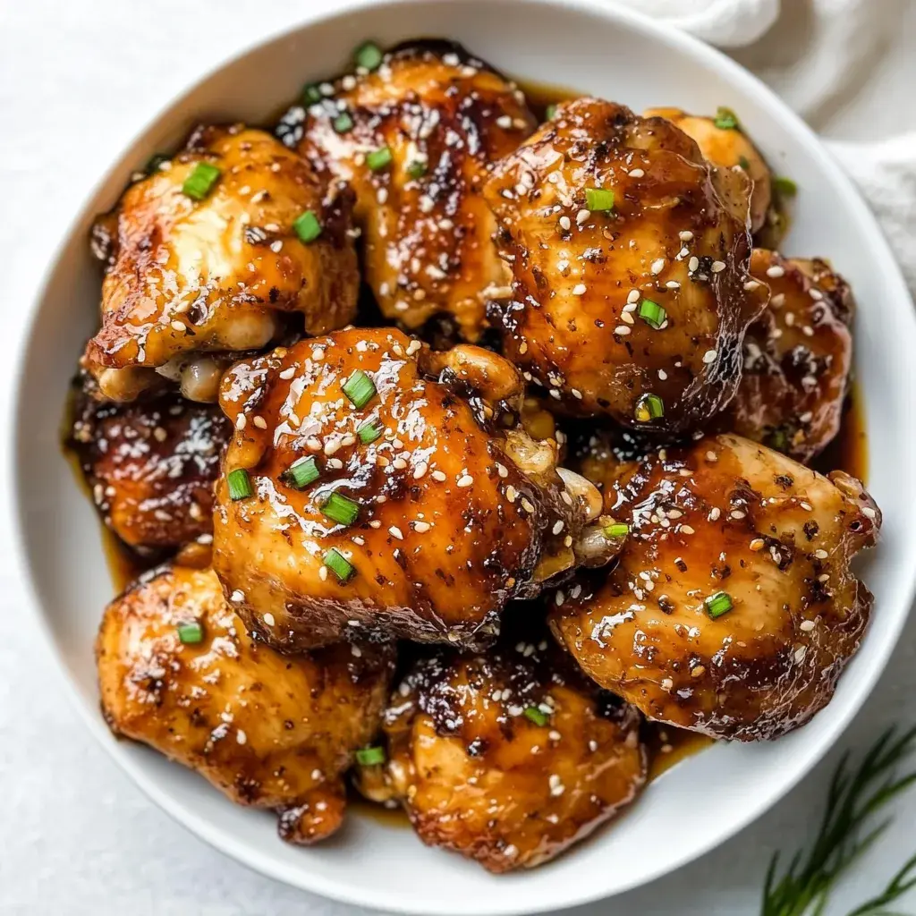 Glazed honey garlic chicken thighs in a white bowl, scattered with sesame seeds and sliced spring onion