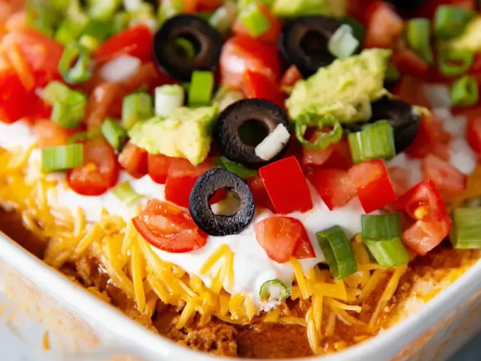 A clear dish of layered dip with refried beans, cheese, sour cream, guacamole, tomato, black olives and green onion in distinct bands