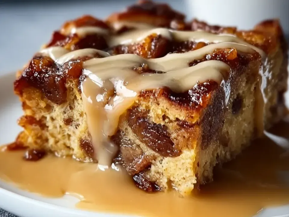 A square-cut slab of raisin-studded snack bread with a caramelised top and soft crumb, drizzled with a pale butter-rum sauce pooling on the plate