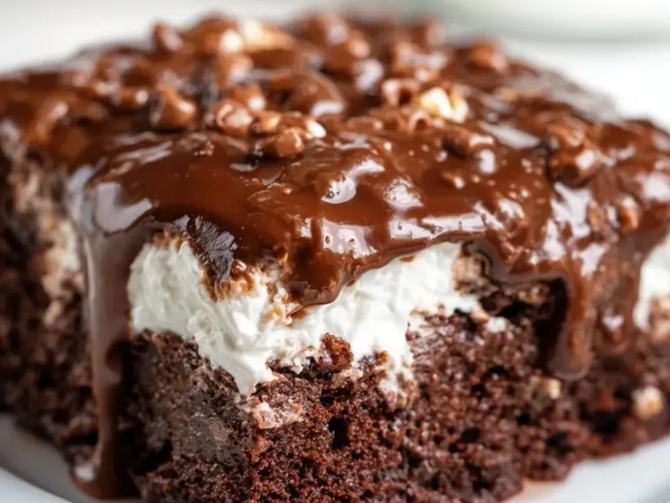 A square of dark chocolate sheet cake under a white marshmallow layer, covered in a glossy poured chocolate frosting studded with chopped pecans
